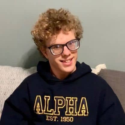Photo of Scott, smiling with blonde curly hair, wearing a blue sweatshirt with "Alpha" written on it.
