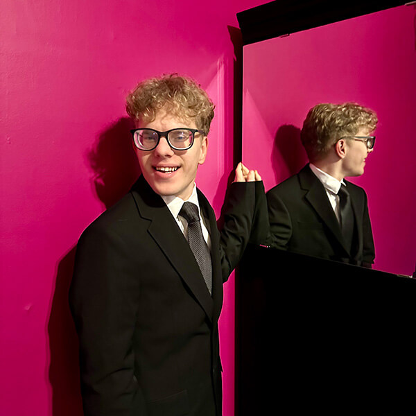 Scott standing next to a mirror, wearing a black suit in a very pink room.